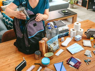 A woman packs an emergency go-bag at a table spread with water, batteries and first-aid supplies