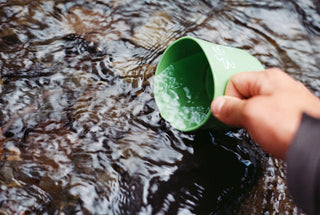 A hand dipping a green cup into a flowing stream
