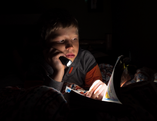 A boy reading a book by flashlight in a darkened room