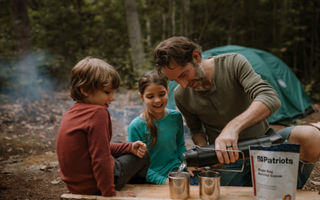 A man pouring a drink for two children at a picnic table beside a 4Patriots pouch
