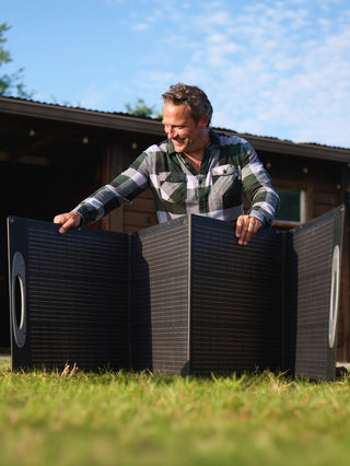 Smiling man in a plaid shirt unfolds the 200-Watt Portable Folding Solar Panel on a grassy farm yard by a barn, off-grid emergency backup solar power
