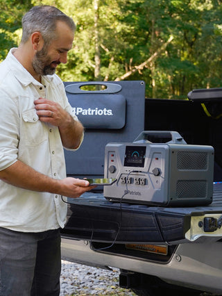 200-Watt Portable Folding Solar Panel behind a Patriot Power Generator 2200X on a truck tailgate, charging a bearded man's phone off-grid in the woods for emergency backup power