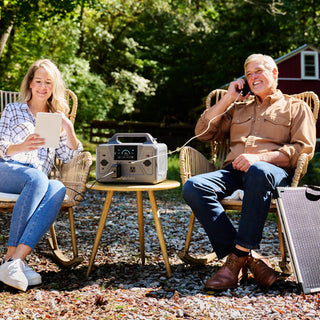 Couple outdoors charging a tablet and phone from the Patriot Power Generator 1200 on a side table, portable solar power station for camping