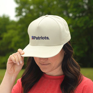 America's 250 Year Anniversary Hat worn by a woman outdoors tipping the brim, cream 4Patriots logo cap against a backdrop of green trees