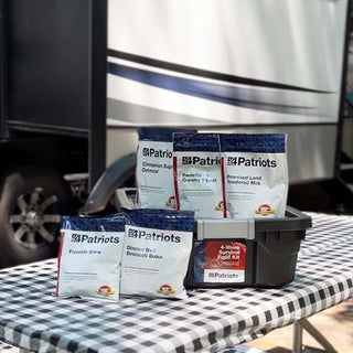 4-Week Survival Food Kit: 4Patriots meal pouches and tote on a picnic table beside an RV, survival food for camping and emergencies