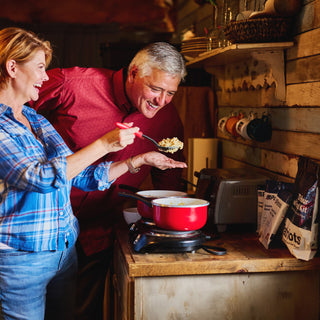 Smiling couple cooking and tasting 72-Hour Emergency Survival Food Kit emergency meals on a hot plate in a cabin kitchen with food pouches nearby