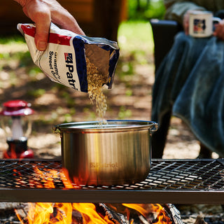 Hand pouring 72-Hour Emergency Survival Food Kit oatmeal into a pot over a campfire, cooking freeze-dried camping food outdoors