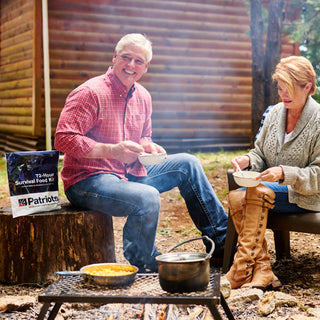 Couple eating bowls of hot 72-Hour Emergency Survival Food Kit meals by a campfire and cabin with a food pouch beside them, camping food