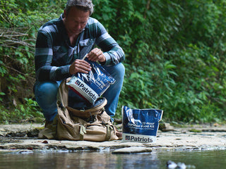 Man opening a 72-Hour Emergency Survival Food Kit pouch beside a backpack and second kit by a stream, freeze-dried survival food