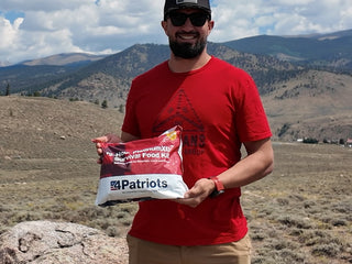 Smiling man in the mountains holding a 72-Hour PlatinumXL Emergency Survival Food Kit pouch outdoors, camping food for backcountry trips