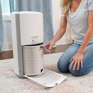 Woman kneeling on the floor next to a 4Patriots air purifier with the bottom panel open, pulling out the cylindrical HEPA replacement filter for maintenance