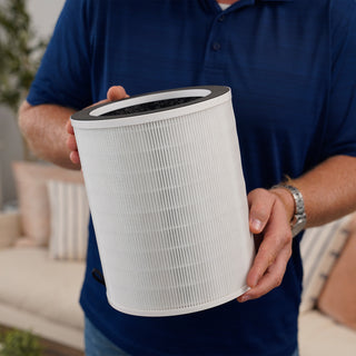 Man in a blue polo shirt holding a cylindrical 4Patriots air purifier H13 HEPA replacement filter with activated carbon top layer in a living room setting