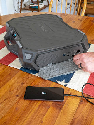 Patriot Power Solar Generator AlphaCase Elite on a table with open port panel of USB and AC outlets charging a smartphone, solar phone charger
