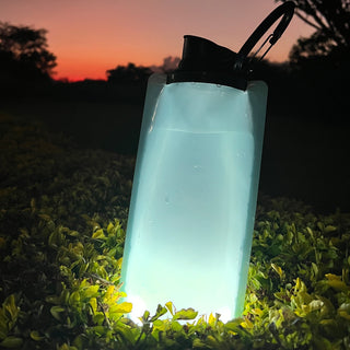 Aqua-Bright 2-in-1 Water Bottle & Lantern glowing as a lantern in grass at dusk, doubling as an emergency light and survival gear for camping