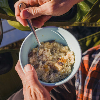 Hands holding a bowl of Country Griddle Breakfast Kit oatmeal with raisins outdoors, a warm breakfast on the trail