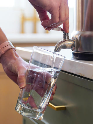 Close-up of a hand filling a glass from the stainless spigot of the Patriot Pure Ultimate Water Filtration System on a kitchen counter.