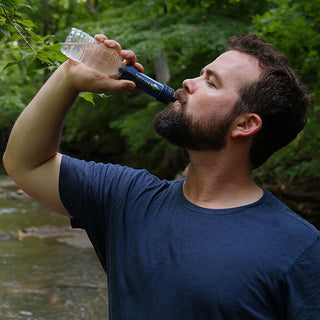 Man drinks from a plastic bottle fitted with the Patriot Pure Personal Water Filter Straw beside a wooded creek.