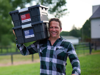 Smiling man carries two stacked 3-Month Survival Food Kit totes from the Family Favorites Emergency Survival Food Package on his shoulder outdoors.
