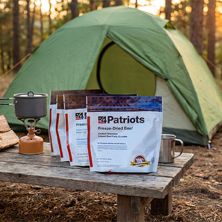 Freeze-Dried Beef and entree pouches from the Family Favorites Emergency Survival Food Package on a picnic table with a camp stove and tent at sunrise.