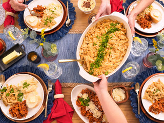 Overhead flat lay of hands passing a noodle casserole at a table set with meals from the Family Favorites Emergency Survival Food Package.