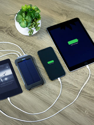 Patriot Power Cell-CX Portable Solar Power Bank on a wood table charging an e-reader, smartphone and tablet at once, all shown at 100%, portable solar power bank for off-grid emergency backup power
