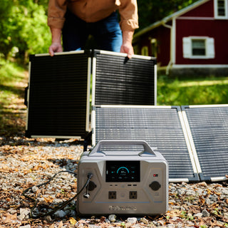 Gold Reserve Emergency Survival Package: man setting up two folding solar panels behind the Patriot Power Generator 1200 outside a red cabin, off-grid power