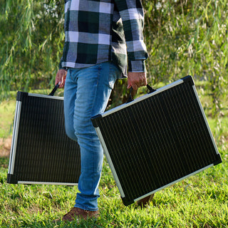 Gold Reserve Emergency Survival Package: man carrying two foldable 100W solar panels by their handles across a grassy farm, portable off-grid power