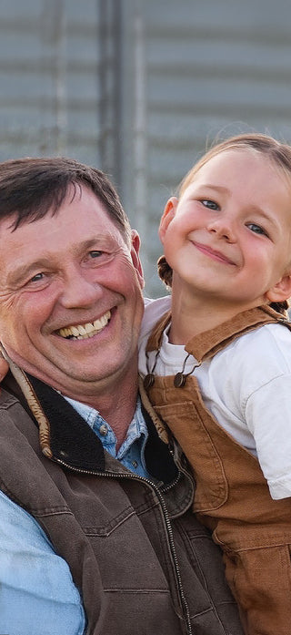 Smiling man in a work vest embracing a young child in overalls outdoors by a metal grain silo, a 4Patriots lifestyle image conveying family disaster preparedness and self-reliance