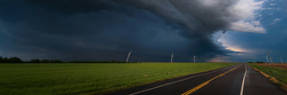 Ominous storm front rolling over green fields, wind turbines and a wet country road, a wide severe-weather scene evoking blackouts and the need for emergency backup power