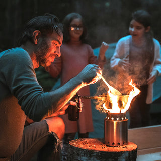 StarFire Portable Camp Stove burning bright as a dad lights it at dusk while his daughters watch, a wood-fueled off-grid camp stove for camping and emergency cooking