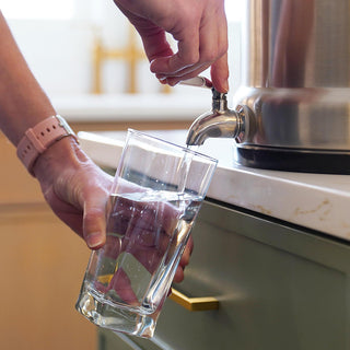 Patriot Pure Ultimate Water Filtration System dispensing clean water into a glass on a kitchen counter, a countertop gravity water filter for emergency preparedness