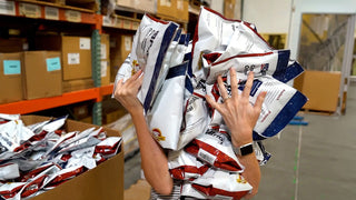 4Patriots survival food pouches carried by a woman past a warehouse pallet rack, a 4-Week Survival Food Kit buy-one-get-one deal for emergency food storage