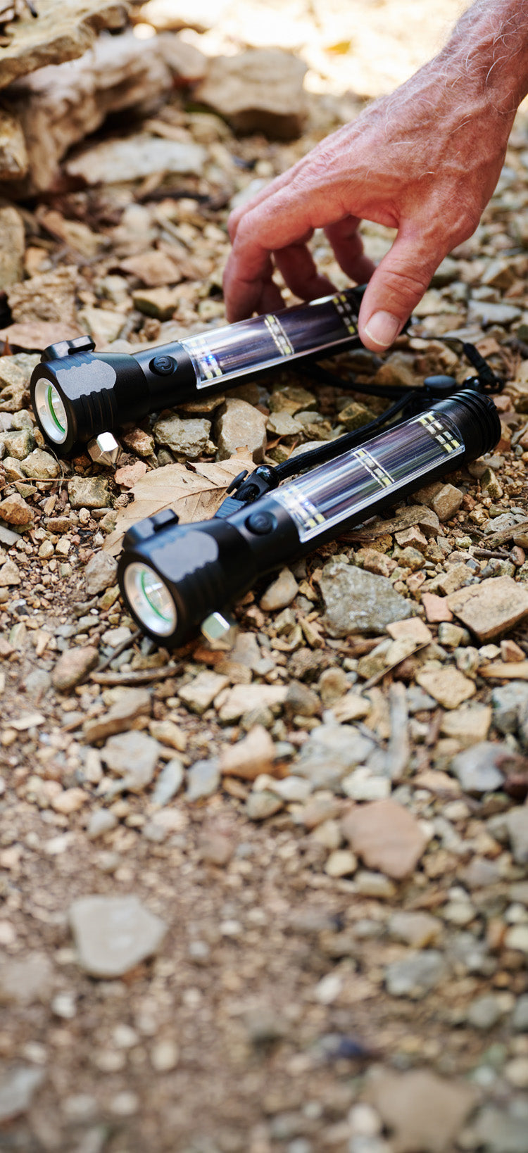 Translation missing: en.HaloXT Tactical 9-in-1 Solar Flashlight, two rugged units resting on gravel and leaves as a hand reaches down, solar-rechargeable off-grid emergency flashlight for disaster preparedness