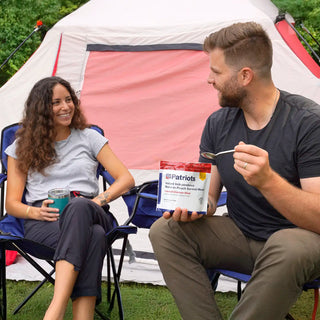 Smiling man and woman in camp chairs by a tent; he holds a 4Patriots Loaded Chicken Stew pouch and a spoon.