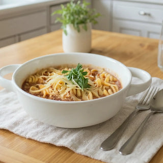 White two-handled bowl of pasta in marinara sauce topped with shredded cheese and parsley, with a fork and spoon on a linen cloth
