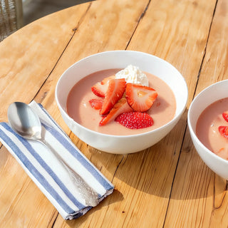 White bowl of strawberries and cream with sliced strawberries and whipped cream, beside a spoon on a striped napkin on a wooden table