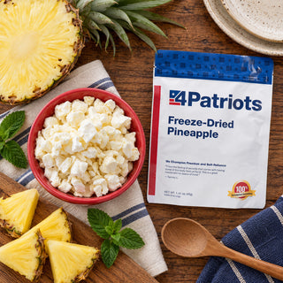 Freeze-Dried Pineapple Pouch flatlay with a red bowl of pineapple bites, a halved pineapple, fresh wedges, mint and a wooden spoon, emergency food supply