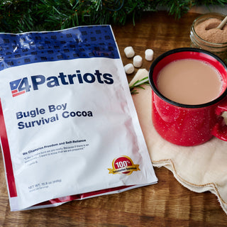 4Patriots Bugle Boy Survival Cocoa Pouch beside a red camp mug of hot cocoa with marshmallows and a jar of cocoa powder, comforting survival food flatlay
