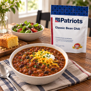 Classic Bean Chili Pouch behind a bowl of bean chili with shredded cheddar and sour cream, plus cornbread and a garden salad on a table, survival meals