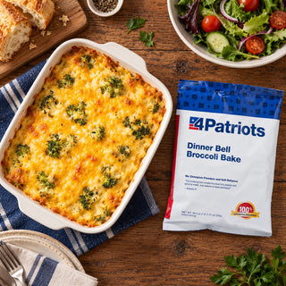Overhead view of Dinner Bell Broccoli Bake Pouch beside a golden broccoli and cheese casserole, crusty bread and tomato cucumber salad, emergency food supply