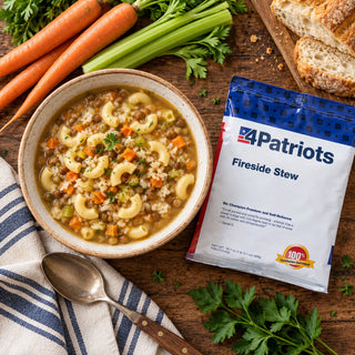 Top-down Fireside Stew Pouch flatlay with a bowl of macaroni lentil stew, whole carrots and celery, sliced bread and a vintage spoon on wood, emergency food supply