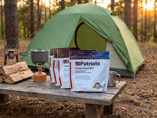 Gold Medallion All-Meat Emergency Survival Kit freeze-dried beef pouches at a campsite with tent and camp stove, portable emergency survival food for camping and preparedness