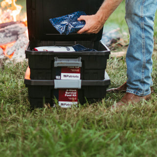 4-Week Survival Food Kit buy-one-get-one: two black totes by a campfire as a man pulls a food pouch from the top kit, ready-to-eat survival food for off-grid long-term food storage