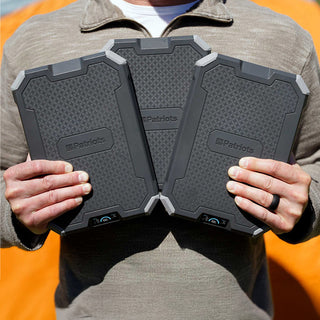 Person holding a 3-pack of Patriot Power Voyager XL solar power banks, three rugged black portable chargers fanned out, off-grid portable solar power for emergency preparedness