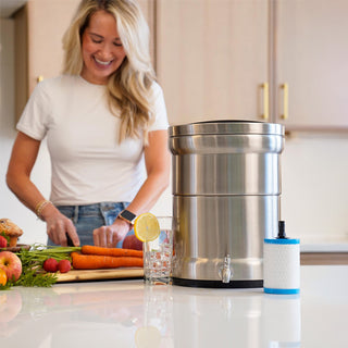Patriot Pure Ultimate Water Filtration System, a stainless steel gravity water filter with spigot and spare cartridge on a kitchen counter as a woman chops vegetables for preparedness