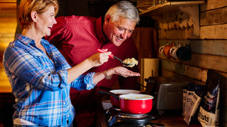 Smiling older couple cooking 4Patriots survival food on a small stove in a rustic cabin kitchen, the woman offering a taste, long-term food storage and emergency meals for disaster preparedness