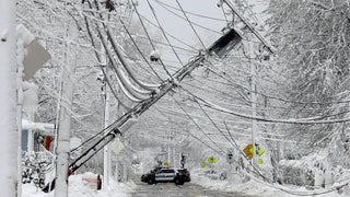 Heavy snowstorm with a leaning utility pole and downed power lines over a snow-covered street as a police SUV blocks the road, a winter blackout scene showing the need for emergency backup power