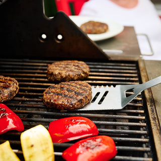 Four 4Patriots Black Bean Burger Mix patties grilling alongside red bell pepper halves and yellow squash slices on a gas grill, with a metal spatula and a plated burger in the background