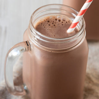 Glass mason jar mug filled with a creamy 4Patriots Survival Shake Chocolate drink topped with foam and a red and white striped straw, with a second jar visible in the background