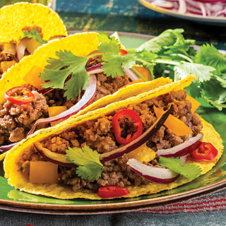 Two yellow corn taco shells filled with 4Patriots Taco Flavored Plant Based Protein, topped with red chili slices, red onion, yellow bell pepper, and fresh cilantro, served on a green plate with additional tacos in the background